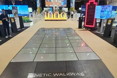 Chodnik z płyt **piezoelektrycznych** na targach, generujący energię z kroków. W tle wielkie ekrany i logo "INNO".