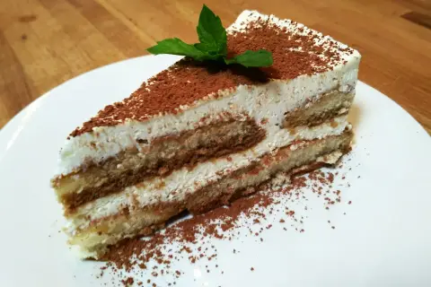 Pyszny kawałek najprostszego tiramisu, posypany kakao i udekorowany listkiem mięty, na białym talerzu.