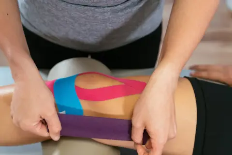 Terapeuta zakłada kolorowe tejpy na kolano, pokazując, co to są tejpy i jak mogą wspierać mięśnie i stawy.
