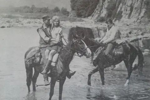 Wilcze echa obsada: para na koniu w rzece, obok drugi jeździec.