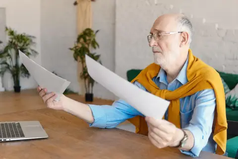 Starszy mężczyzna w okularach przegląda dokumenty przy biurku z laptopem.