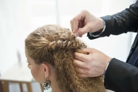 Hårstylist flätar en del av en kvinnas lockiga hår. En bra frisyr kan ge inspiration för hur man får lockigt hår.