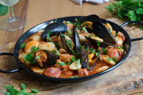 Un plat de moules au chorizo, garni de persil frais, servi dans une poêle noire sur une planche en bois.