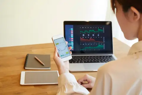 Frau analysiert Aktienkurse auf Laptop und Smartphone. Sie plant, wie sie ihre Aktiengewinne versteuern kann, während sie Notizen macht.