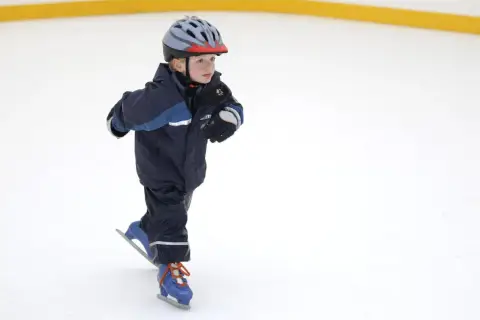 Bambino con casco e tuta blu pratica il pattinaggio sul ghiaccio, con i pattini azzurri e lacci arancioni.