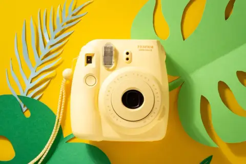 Żółty aparat natychmiastowy Fujifilm Instax Mini 8 na tle żółtym z zielonymi i niebieskimi liśćmi. Idealny do wakacyjnych zdjęć, często pojawia się w rankingach aparatów natychmiastowych.