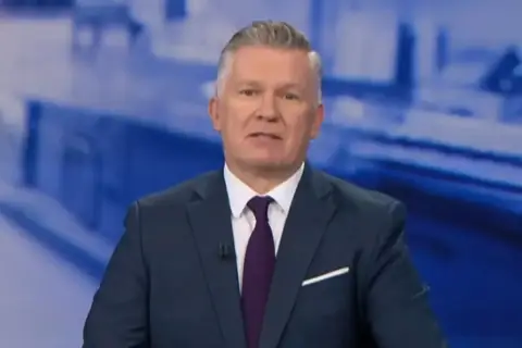 Jeden z prezenterów Fakty TVN w granatowym garniturze i bordowym krawacie, na tle niebieskiej grafiki.