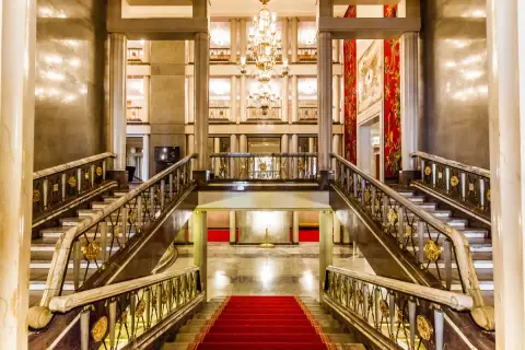 Elegancki foyer z marmurowymi schodami, złotymi balustradami i kryształowym żyrandolem. Czerwony dywan prowadzi do przestronnego holu.