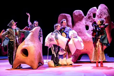 Aktorzy w barwnych strojach, niosący poduszki, na tle scenografii przypominającej gigantyczne, różowe formacje. To fragment spektaklu z repertuaru teatru lalki.