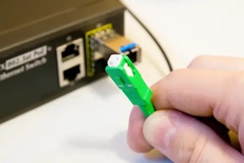 Dłoń trzyma zieloną wtyczkę światłowodową. Obok widać przełącznik Ethernet. Tak sprawdzisz, czy jest światłowód.