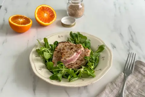 Tonno in padella con crosta di sesamo su letto di insalata fresca, accompagnato da spicchi d'arancia.