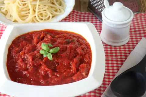 Un tazón blanco lleno de salsa marinara casera, decorada con albahaca fresca, junto a espaguetis cocidos.