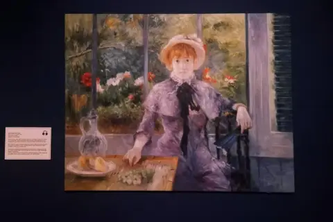 Auguste Renoir dzieła: portret młodej kobiety w kapeluszu, siedzącej przy stole z owocami i dzbankiem, w otoczeniu kwitnącego ogrodu.