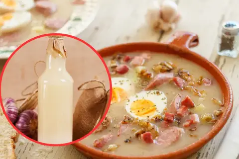 Miseczka żurku z jajkiem i kiełbasą, obok butelka zakwasu, czosnek i chleb.