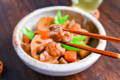 A close-up of a bowl of chikuzenni recipe, with chopsticks lifting a piece of chicken. The dish contains chicken, lotus root, carrots, and green beans.