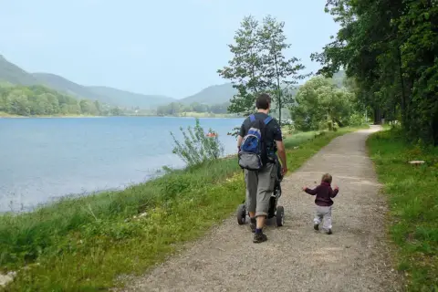 Vater schiebt Kinderwagen am Happurger Stausee entlang, während das Kind vorausläuft. Herrliche Kulisse für eine Wanderung.