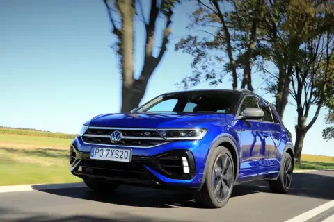 Niebieski Volkswagen T-Roc R pędzi po wiejskiej drodze, otoczony drzewami i polami.