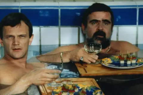 Dwóch mężczyzn w wannie, jeden z wąsami, pije wino i je przekąski. Scena z filmu "Kiler", rok produkcji 1997.