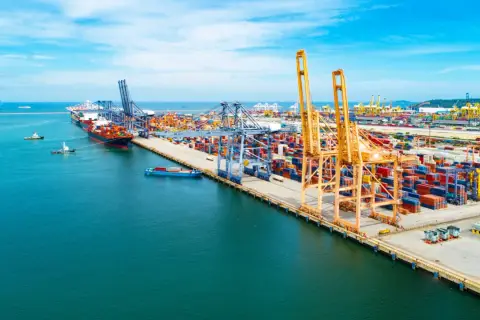 Widok na port kontenerowy z dźwigami, statkami i stosami kontenerów w słoneczny dzień.