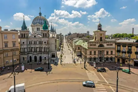 Widok na plac wolności w Łodzi z monumentalną katedrą po lewej i długą ulicą pełną ludzi i samochodów.