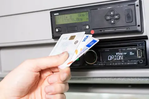 Dłoń wkłada kartę kierowcy do tachografu. Widoczny slot na kartę i ekran urządzenia.