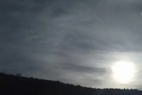 Słońce przebija się przez cienką, włóknistą warstwę cirrostratus chmura, oświetlając sylwetkę lasu na wzgórzu.