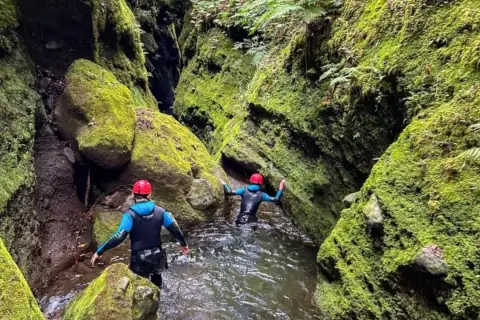 Dwie osoby w kombinezonach i kaskach przemierzają wąski, porośnięty mchem kanion. To właśnie jest canyoning!
