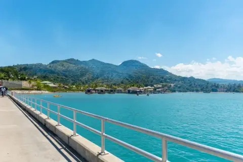 Un muelle con barandilla metálica conduce a un pueblo sobre el agua con montañas verdes de fondo. Ideal para excursiones en Puerto Plata.