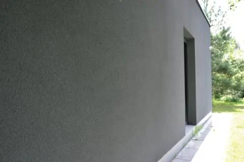 Szara, chropowata elewacja budynku z ciemnym oknem i zielenią w tle.