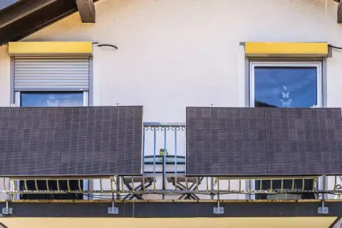 Dwa panele fotowoltaika balkonowa na balustradzie, obok okien z roletami i kwitnących donic.