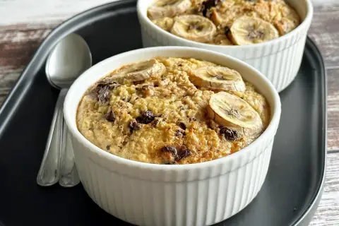 Dwie porcje owsianki z bananem i kawałkami czekolady w białych kokilkach na czarnej tacy. Idealne na słodkie śniadanie.