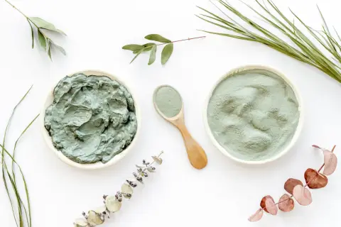 Dos recipientes con arcilla verde, uno en polvo y otro en pasta, junto a una cuchara de madera y ramas secas. Descubre las arcilla verde propiedades.