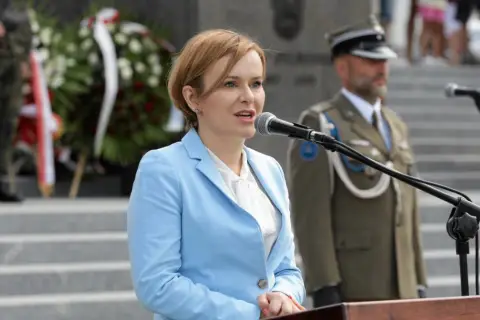 Anna Krupka w błękitnej marynarce przemawia przy mikrofonie. W tle żołnierze i wieńce. Ile milionów złotych oszczędności wykazała w oświadczeniu?