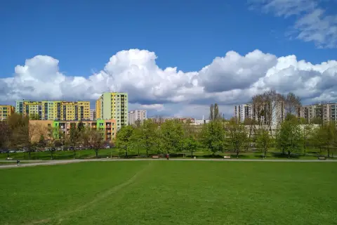 Zielona przestrzeń w parku Górczewska, z kolorowymi blokami mieszkalnymi w tle i białymi chmurami na błękitnym niebie.