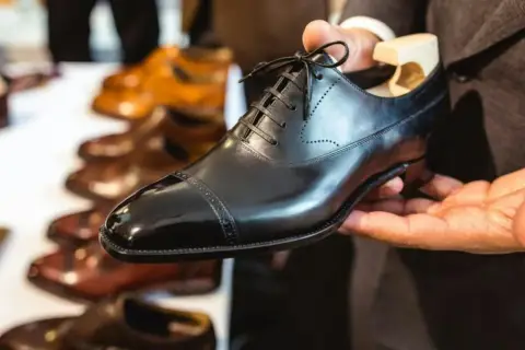Main d'un homme tenant une chaussure noire brillante, prête à nourrir le cuir. D'autres chaussures en cuir sont alignées en arrière-plan.