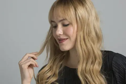 Mujer rubia con cabello ondulado y flequillo, tocando un mechón. A veces, matizar el pelo lo estropea, pero el suyo luce saludable.
