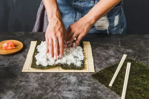 Ręce rozprowadzają ryż na nori, przygotowując zaprawę do sushi. Obok leżą pałeczki i marynowany imbir.