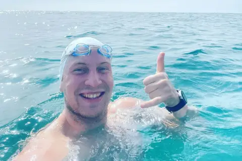 Uśmiechnięty pływak w czepku i goglach, z ręką w geście "hang loose", w trakcie przepłynięcia Bałtyku wpław.