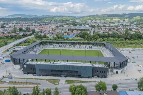 Nowoczesny stadion Sandecji z zieloną murawą i trybunami, otoczony miastem i wzgórzami.