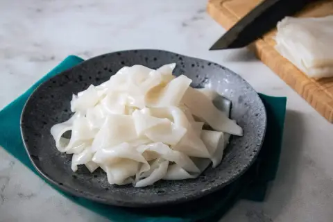 Na talerzu ułożone są białe, szerokie wstążki makaronu, przypominające capelli d'angelo. Obok leży nóż i deska do krojenia.
