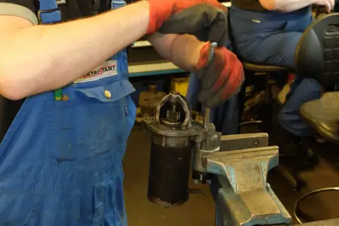 Mechanik w niebieskim kombinezonie i czerwonych rękawicach dokonuje wymiany szczotek w rozruszniku, który jest zamocowany w imadle.