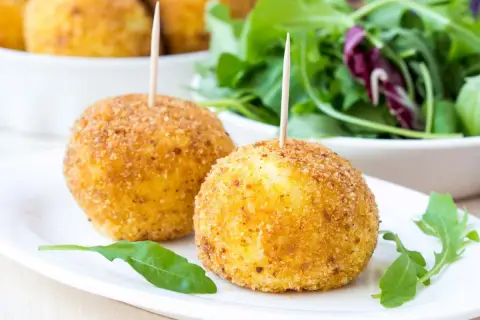 Dwa złociste arancini z wykałaczkami na białym talerzu, obok sałatka.