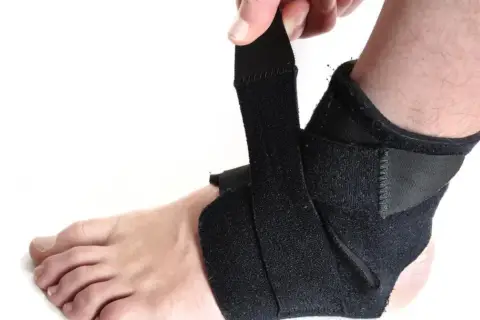 Dłoń zapina czarny stabilizator na kostce. Zapewnia wsparcie i ochronę podczas aktywności.