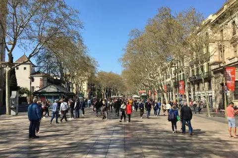 Tłum ludzi spaceruje po słynnej alei **La Rambla w Barcelonie**, otoczonej drzewami i budynkami.