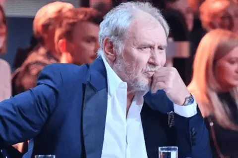 Andrzej Grabowski żyje, siedzi w eleganckim garniturze, zamyślony, z dłonią przy ustach. W tle widać innych ludzi.