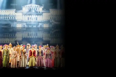 Chór w barwnych strojach historycznych na tle pałacowej scenografii. Sprawdź opera warszawa repertuar!