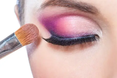 Pędzel do makijażu tworzy wyrazisty **cut crease** na powiece, podkreślając oko intensywnym fioletem i różem.