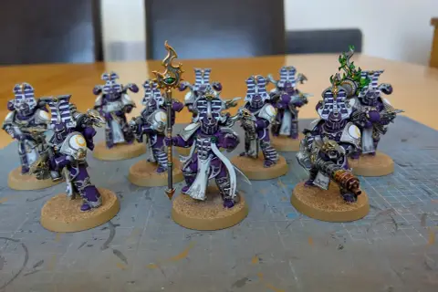 Dziewięciu pomalowanych żołnierzy z Warhammera, w tym jeden tysiąc synów, w fioletowych i srebrnych zbrojach, stoi na piaskowych podstawkach.