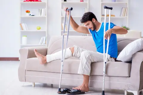 Mężczyzna z zabandażowaną nogą siedzi na kanapie, opierając się o kule. Czeka na rehabilitację po kontuzji, która uniemożliwia mu chodzenie o kulach.