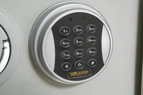 Elektroniczna klawiatura szyfrowa sejfu z przyciskami cyfrowymi i symbolami. W tle widoczne są metalowe tuleje mocujące.